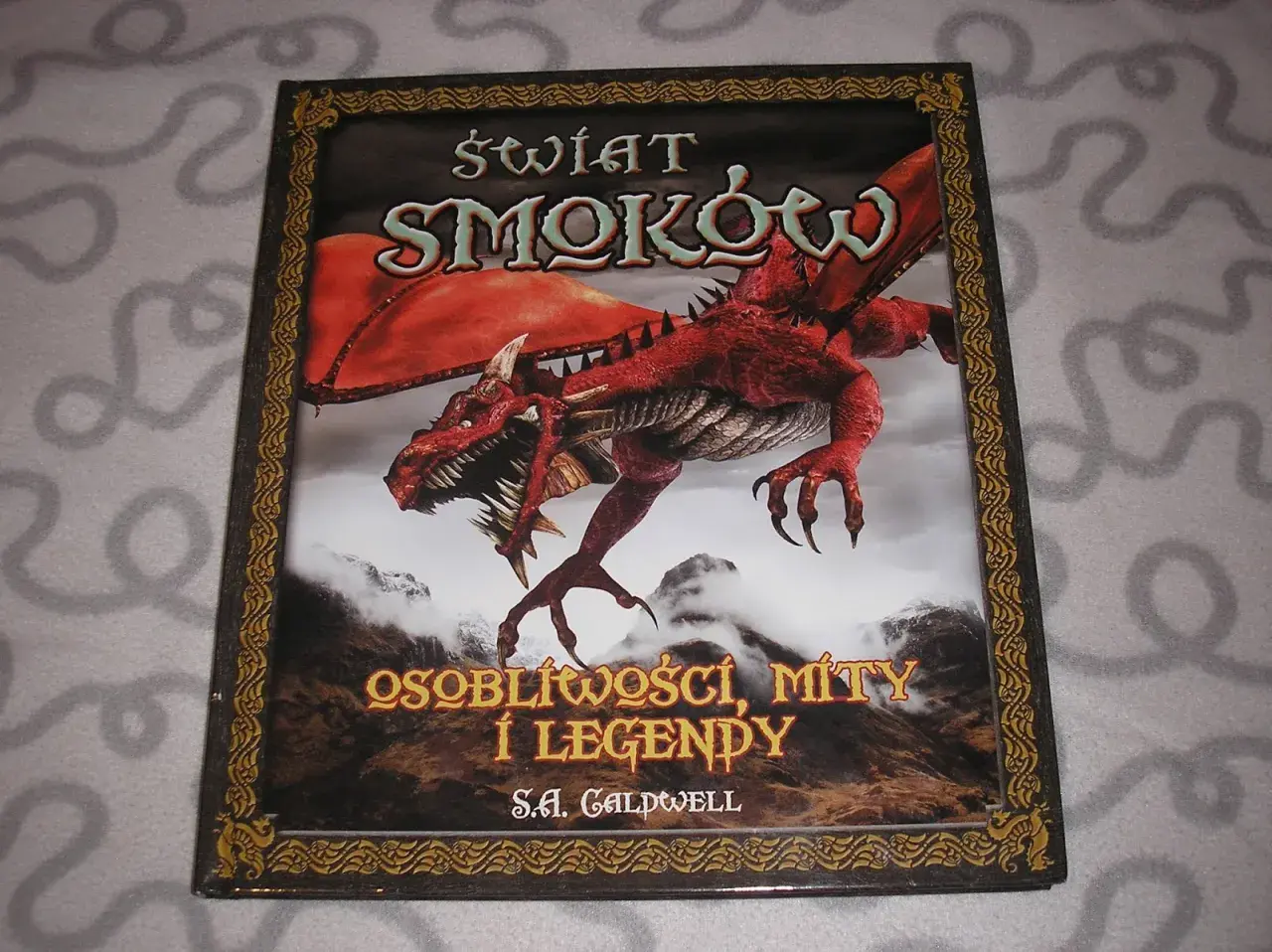 Okładka książki o smokach z czerwonym, groźnym stworzeniem na tle gór. Tytuł: "Świat Smoków: Osobliwości, Mity i Legendy".