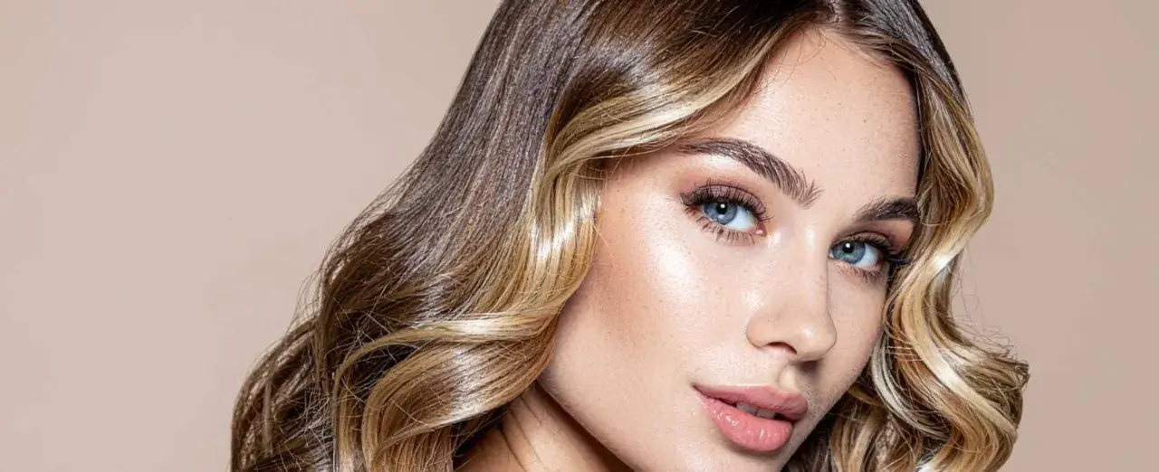 Mujer con cabello castaño y rubio, con un "money piece" que ilumina su rostro. Sus ojos azules y labios rosados resaltan su belleza.