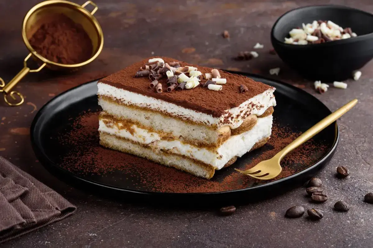Una fetta di tiramisù con philadelphia, cosparso di cacao e riccioli di cioccolato, su un piatto nero con forchetta dorata.