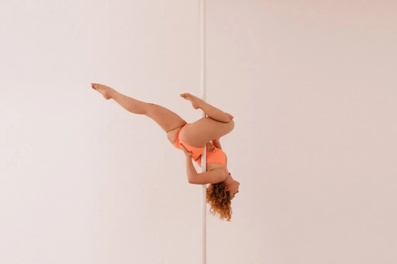 Kobieta w pomarańczowym stroju wykonuje skomplikowaną figurę na pionowym słupku, przypominającą flagę pole dance.