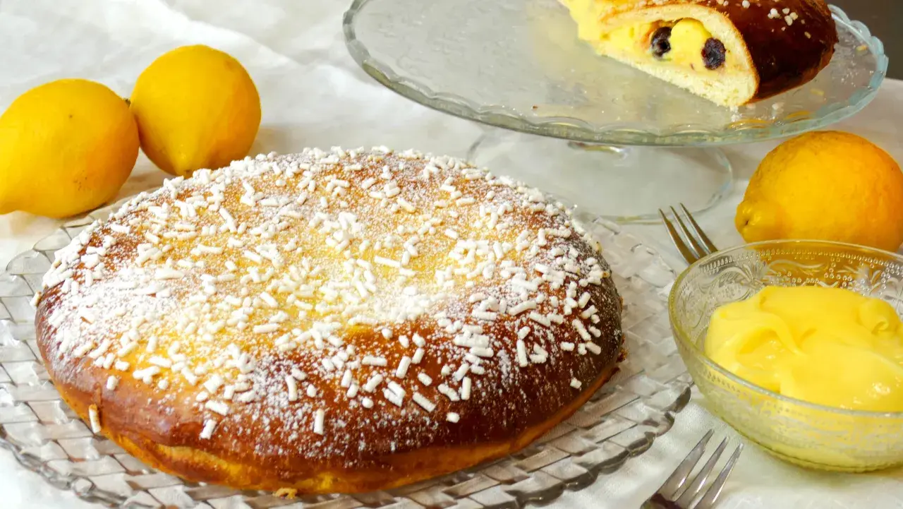 Una deliziosa polacca aversana, cosparsa di zucchero a velo e confettini, accanto a limoni freschi e una ciotola di crema pasticcera.