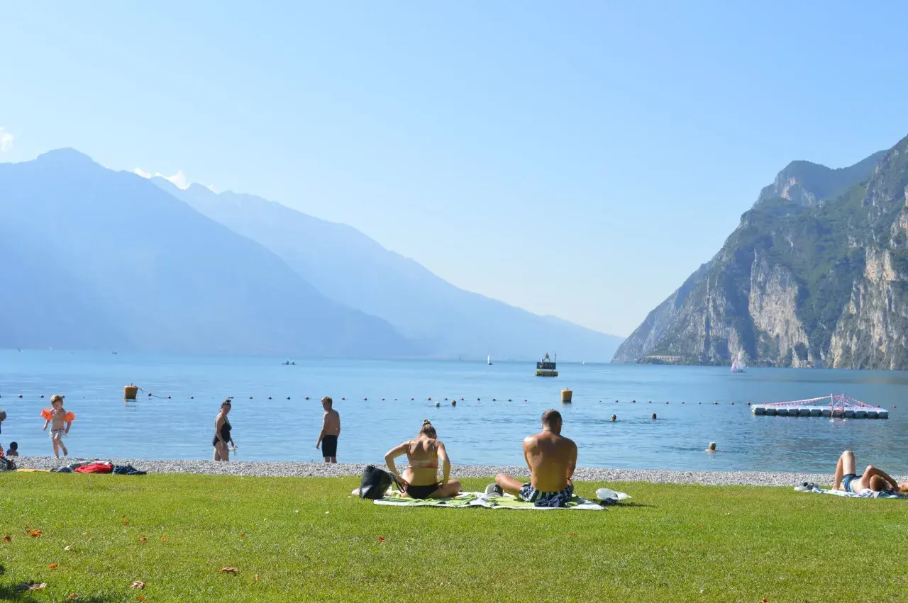 Gente in spiaggia e in acqua, con montagne sullo sfondo, in una giornata di sole sul lago di Garda, forse vicino a Lazise o Bardolino.
