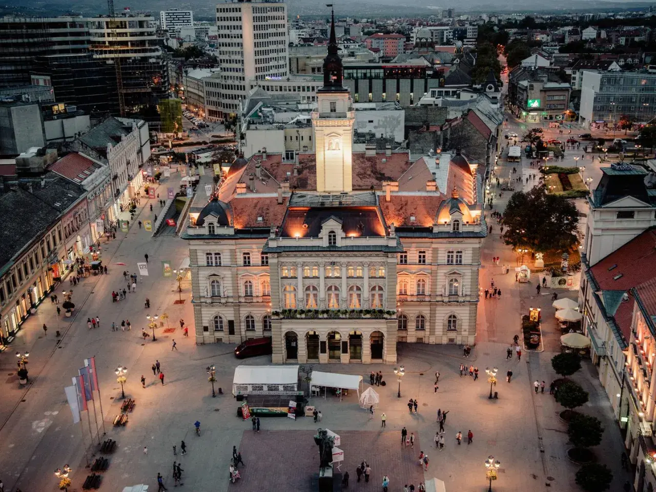 Abendliche Ansicht des Hauptplatzes in Novi Sad, Serbien, mit beleuchteten Gebäuden und Menschen, die spazieren gehen.