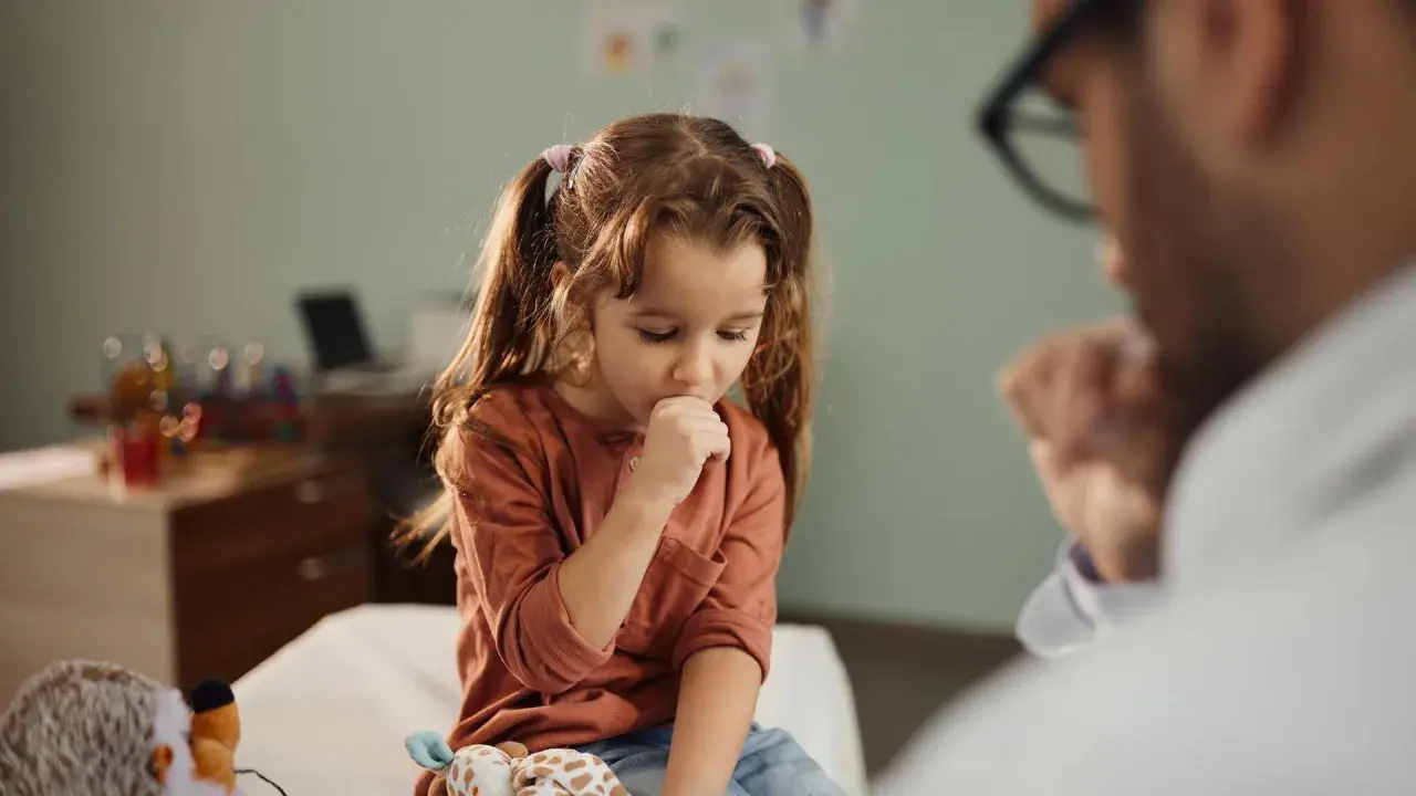 Niña con tos perruna en consulta médica. Un doctor la examina mientras ella se cubre la boca.
