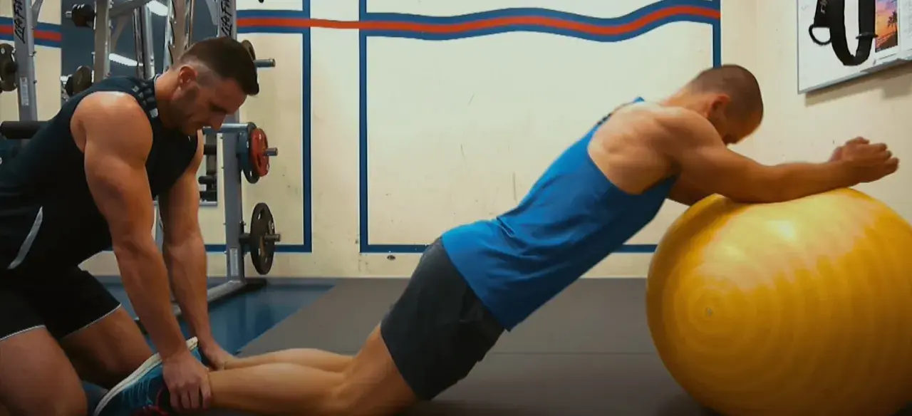 Trener pomaga wykonać ćwiczenie nordic curl, stabilizując nogi klienta opierającego się o dużą, żółtą piłkę gimnastyczną.