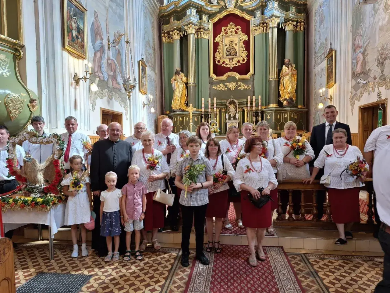 Uroczystość Wniebowzięcia Najświętszej Maryi Panny w kościele. Grupa osób w strojach ludowych i duchowny pozują do zdjęcia.