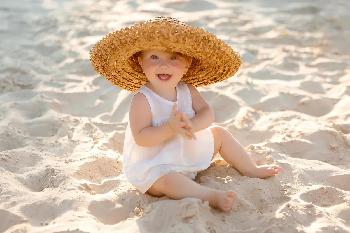 Uśmiechnięte dziecko w słomkowym kapeluszu na plaży. Idealne miejsce, gdzie na wakacje z półrocznym dzieckiem, by cieszyć się słońcem i piaskiem.