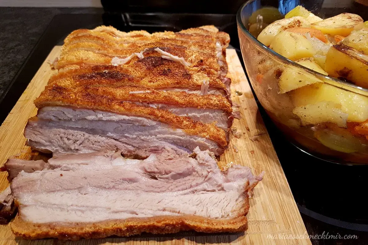 Ein butterzarter Schweinebauch mit knuspriger Kruste liegt auf einem Holzbrett, daneben eine Schüssel mit Bratkartoffeln und Gemüse.