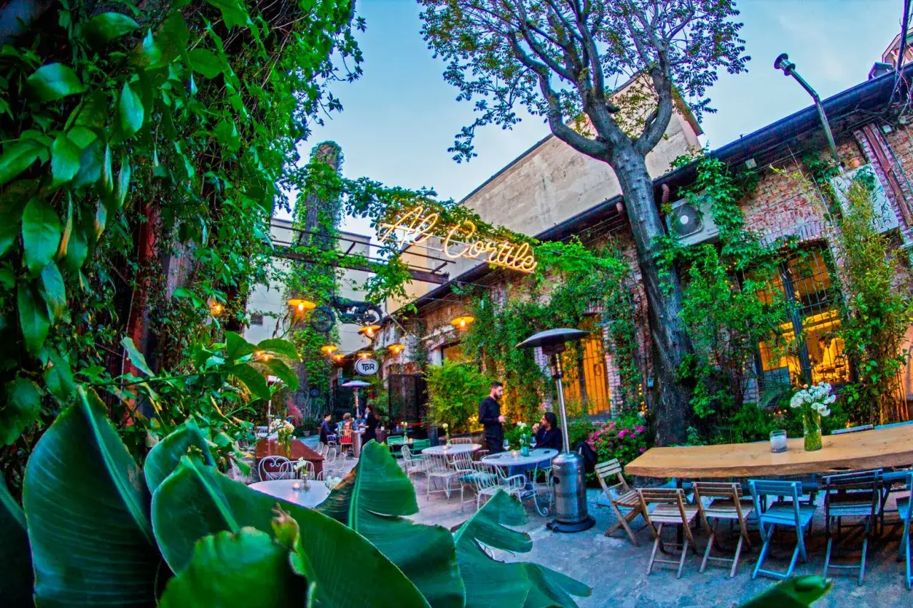 Un cortile rigoglioso ospita aperitivi milanesi particolari. Tavoli all'aperto, luci soffuse e vegetazione creano un'atmosfera magica.