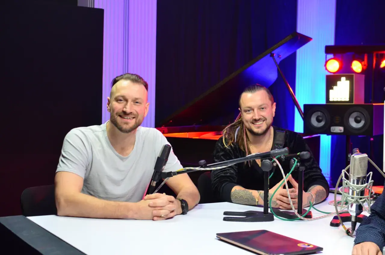 Alek Baron i jego kolega siedzą przy stole w studiu, gotowi do nagrania.