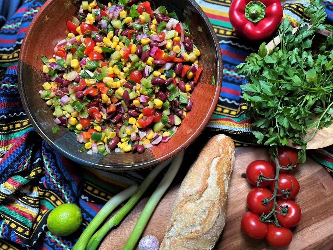 Bunter mexikanischer Salat mit Mais, Bohnen und Gemüse in einer Schüssel, daneben Baguette, Limette und Kräuter.