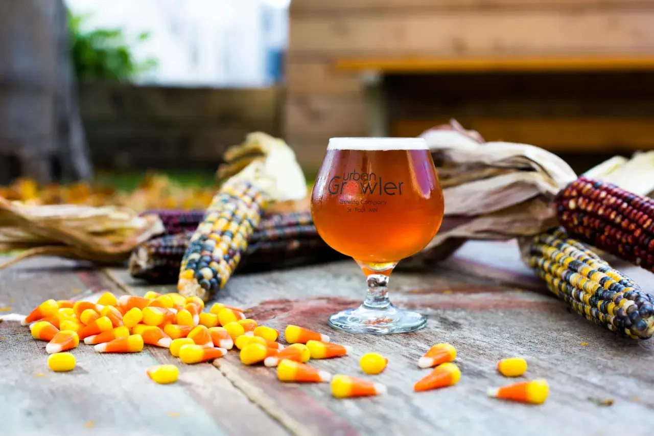 Bicchiere di birra ambrata con logo Urban Growler, circondato da pannocchie colorate e caramelle a forma di mais. Un omaggio autunnale alla birra al mais.
