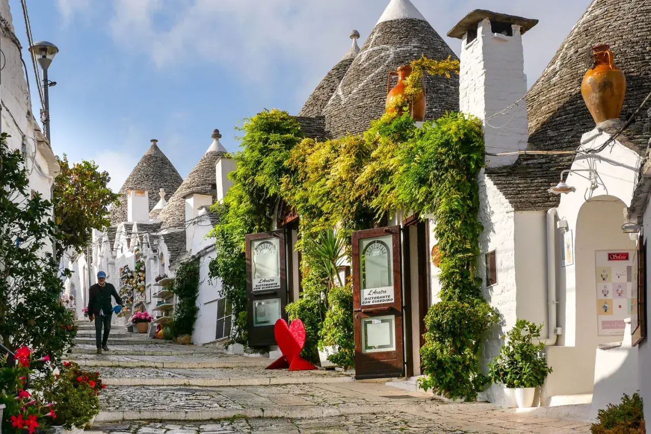 A person walks down a charming street lined with iconic trulli houses, lush greenery, and shops. This picturesque scene makes you wonder, which is the best town to stay in Puglia?