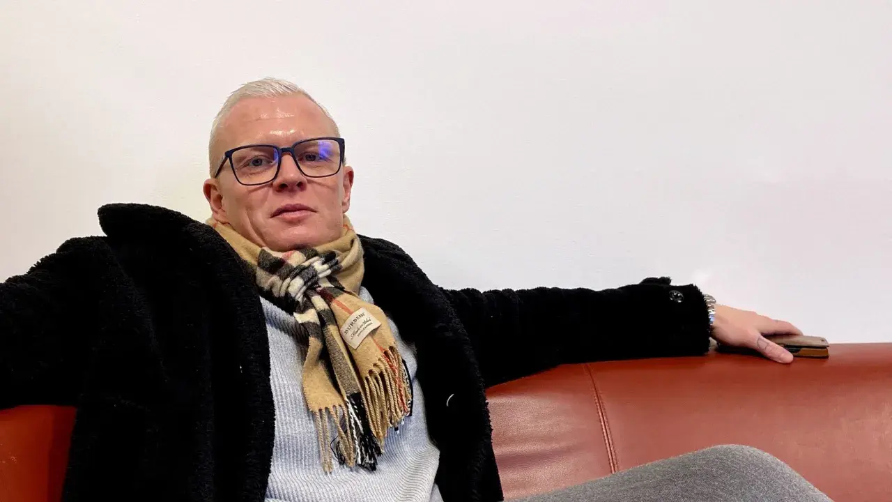 Robinson Alexander, med glasögon och en rutig halsduk, sitter avslappnat på en röd soffa.