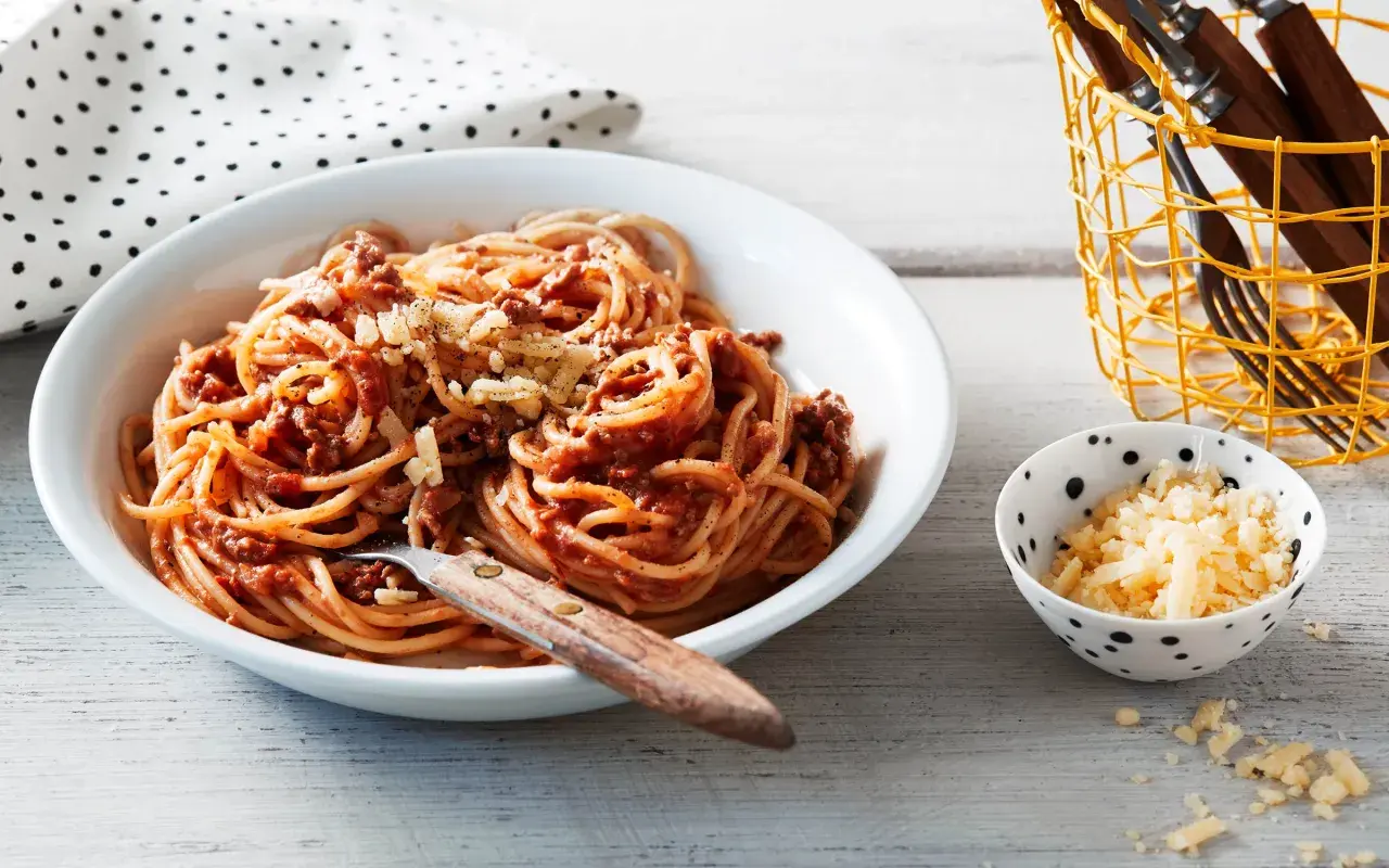 En skål med stuvad spaghetti med köttfärssås och riven ost. En liten skål med mer ost och en korg med bestick står bredvid.