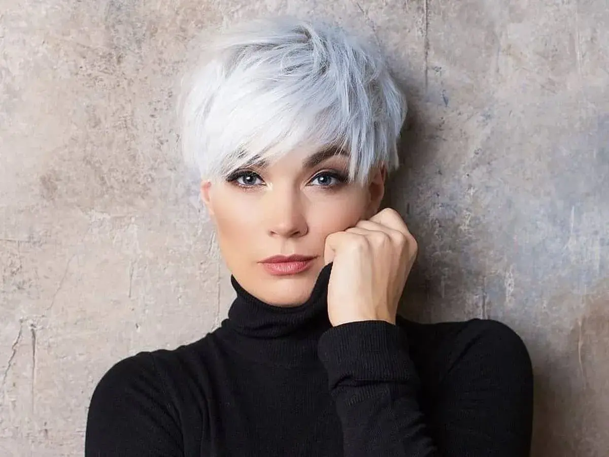 Kobieta z siwymi włosami w stylu pixie bob fryzura, ubrana w czarny golf, opiera dłoń o twarz.