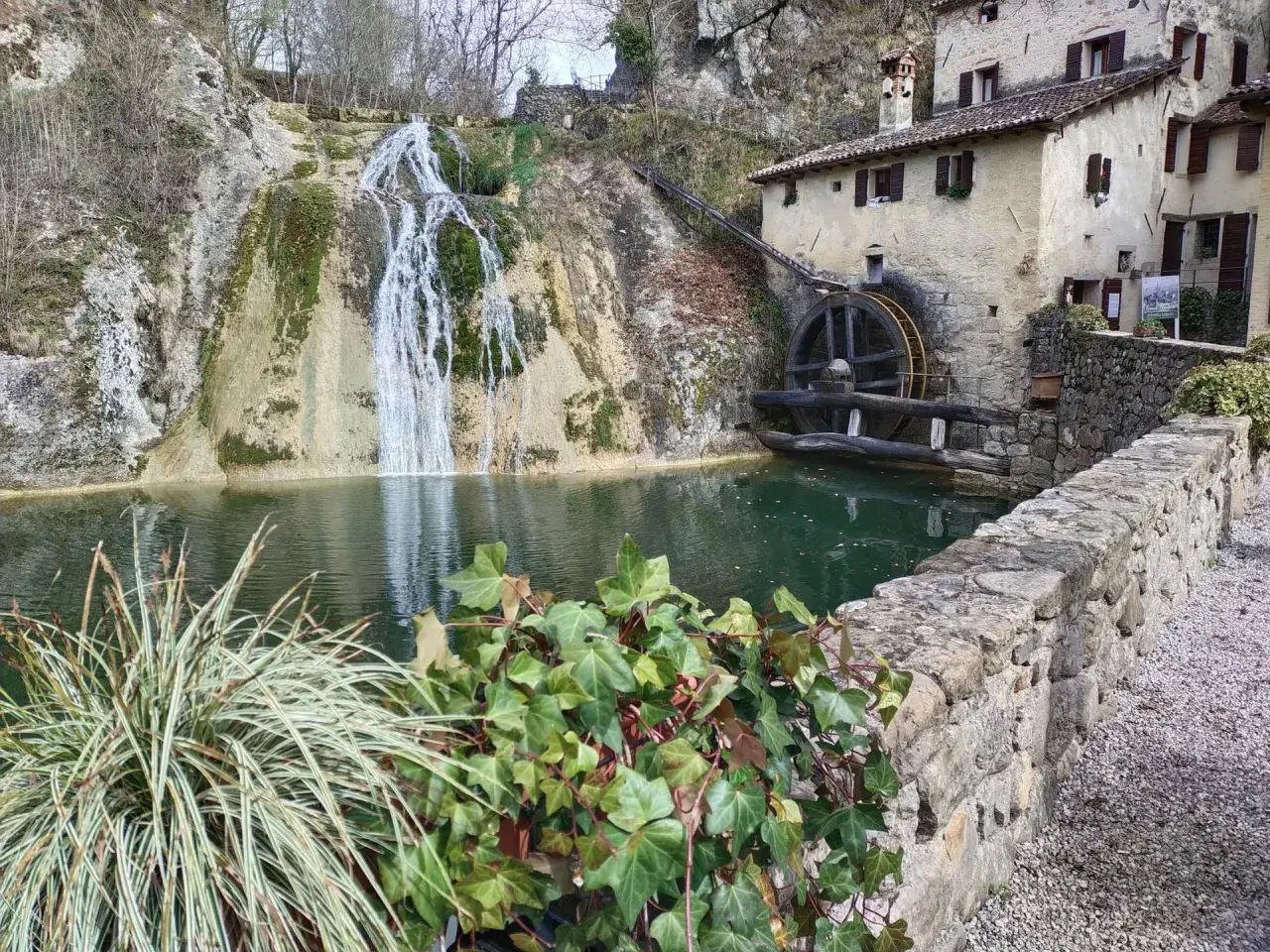 Antico mulino ad acqua accanto a cascate e grotte in Veneto, con un laghetto verde e vegetazione rigogliosa.