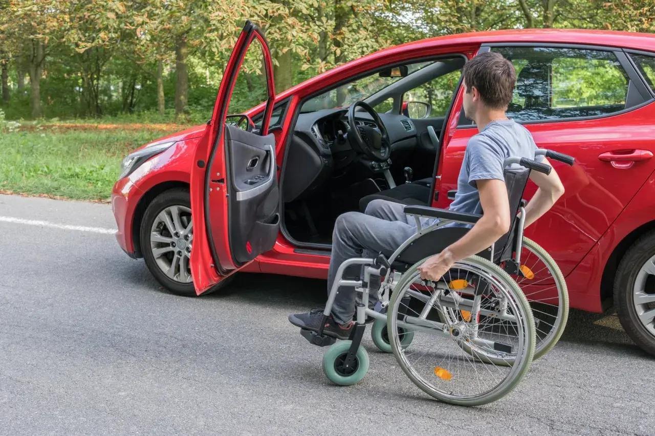 Uomo in carrozzina accanto a un'auto rossa, pronto per il passaggio di proprietà auto disabile decesso.
