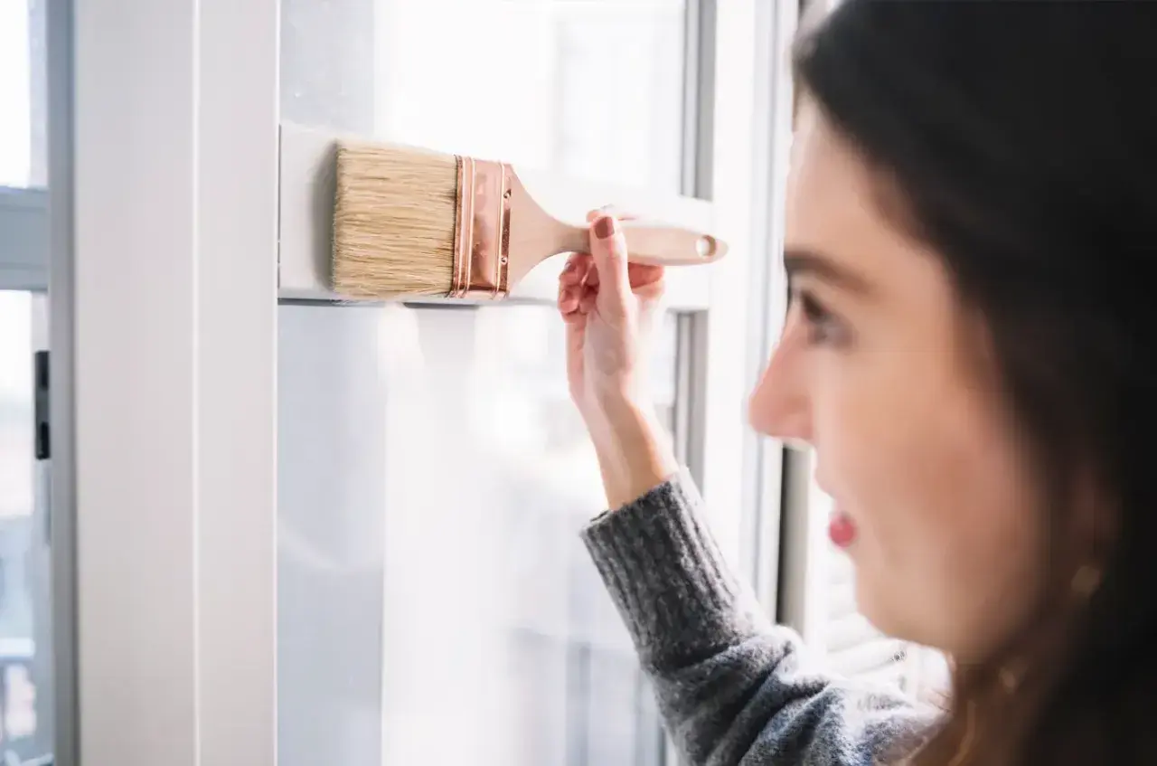Mujer aplica pintura para persianas PVC en un marco blanco.