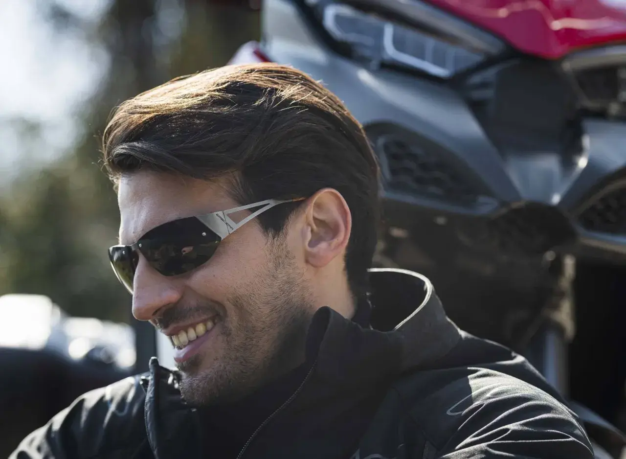 Uomo sorridente con occhiali da moto sportivi, con una moto sullo sfondo.