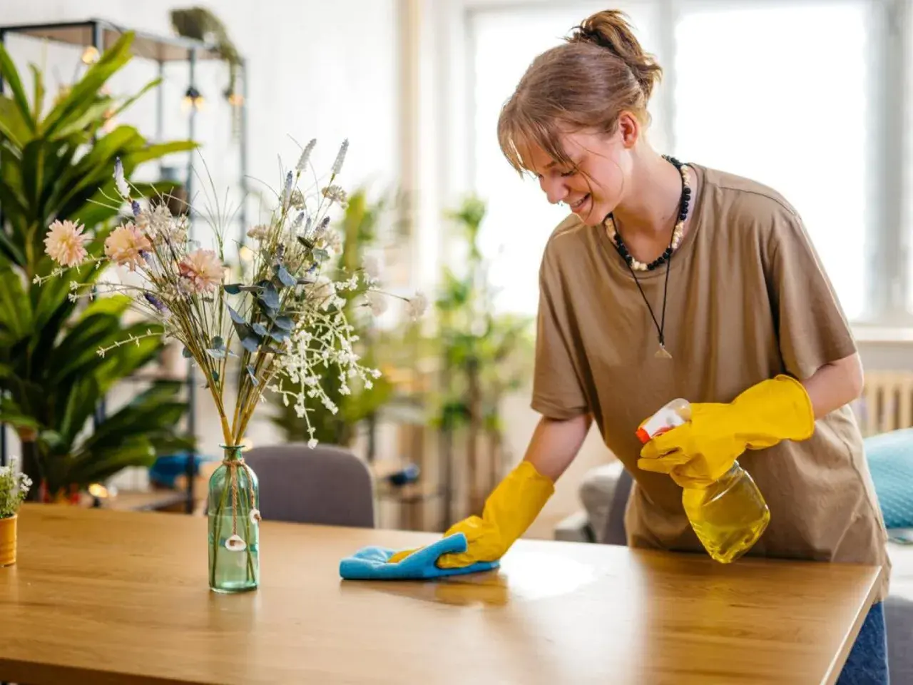 Joven sonríe mientras aprende como limpiar el polvo de los muebles con spray y paño azul.