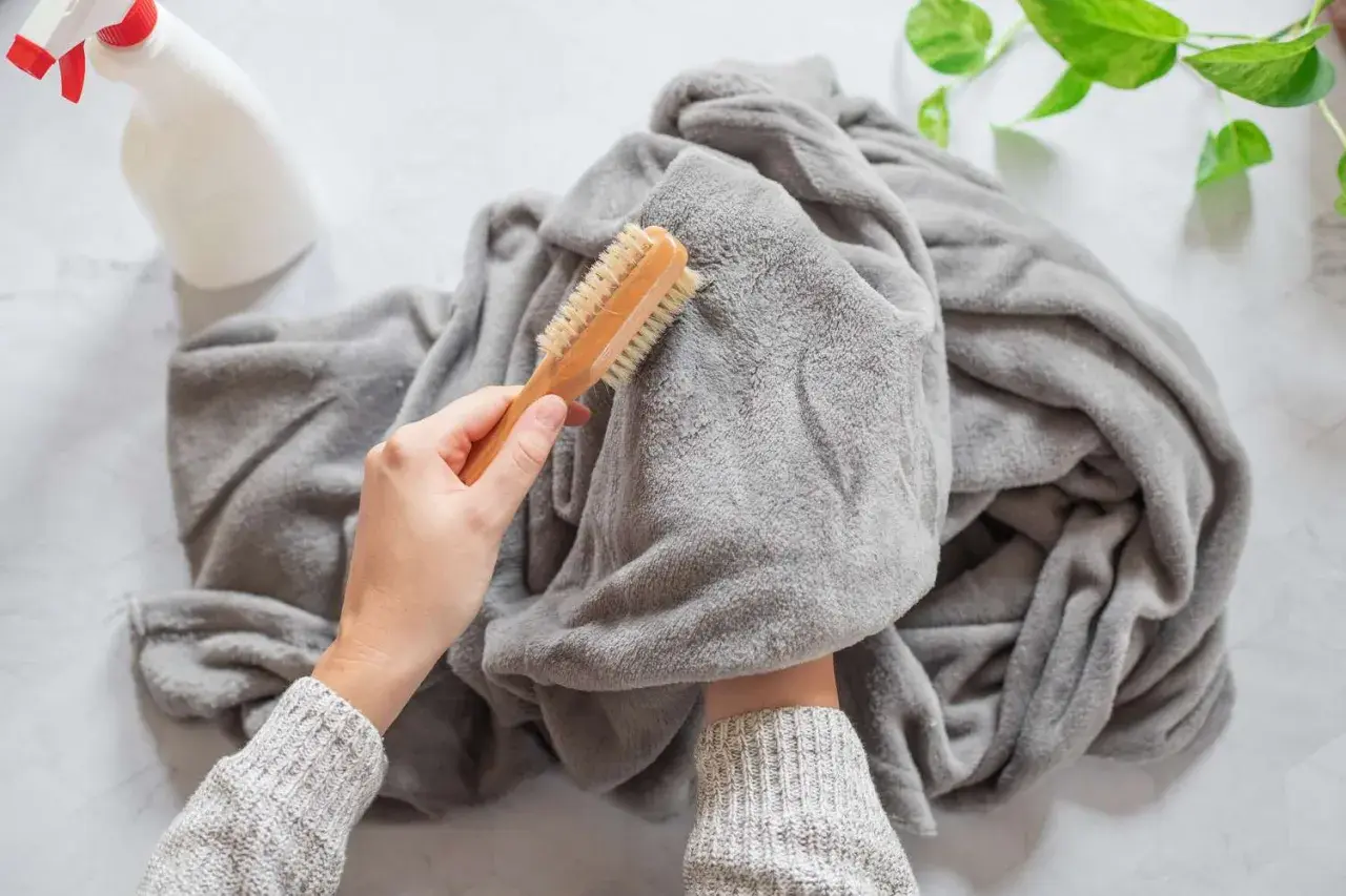 A person is brushing a grey throw blanket, showing how to wash it. A spray bottle is nearby.