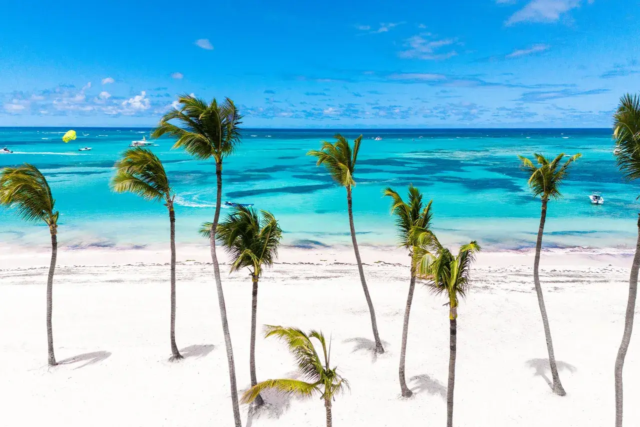 Traumhafter **Urlaub im November** an einem karibischen Strand mit Palmen, weißem Sand und türkisfarbenem Meer. Boote und Parasailing im Hintergrund.