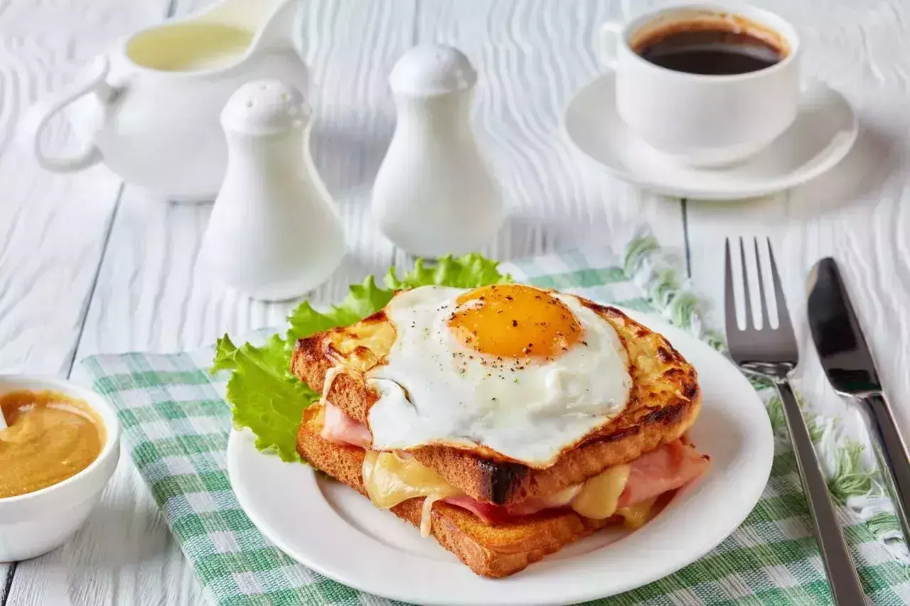 Un délicieux croque monsieur garni d'un œuf au plat, servi avec une tasse de café et une sauce crémeuse.