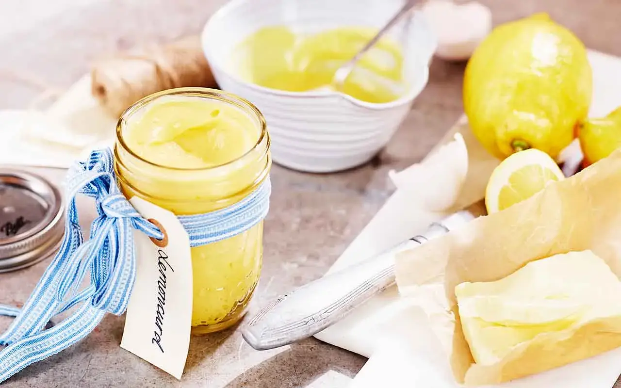 En burk med krämig lemon curd, redo för ett recept. Citroner och smör ligger bredvid.