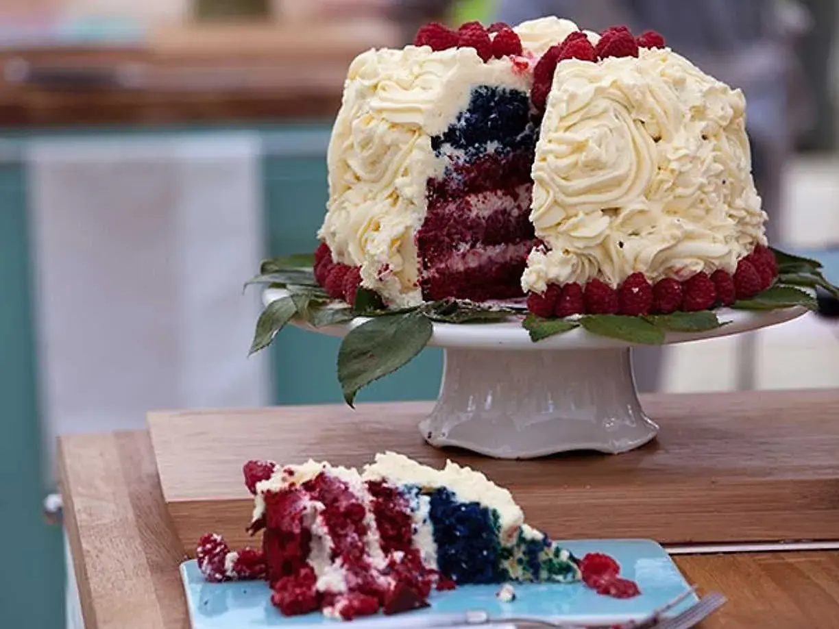 En skivad red velvet cake med vit frosting, hallon och blått inuti. En bit ligger på en blå tallrik.