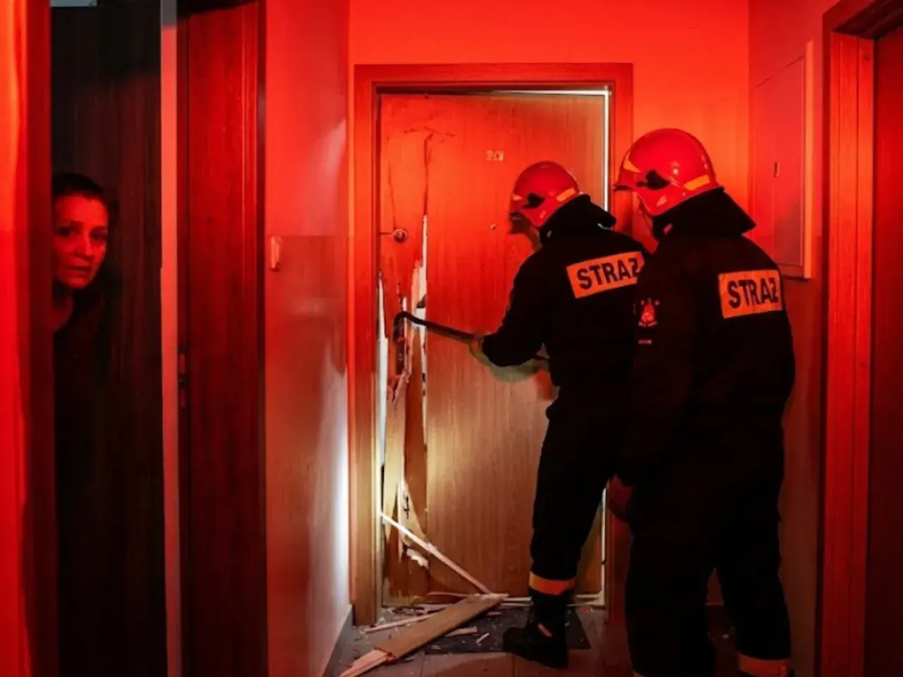 Strażacy wyważają drzwi, gdy kobieta patrzy z niepokojem. Sytuacja, kiedy policja może wyważyć drzwi, by ratować życie lub zapobiec przestępstwu.
