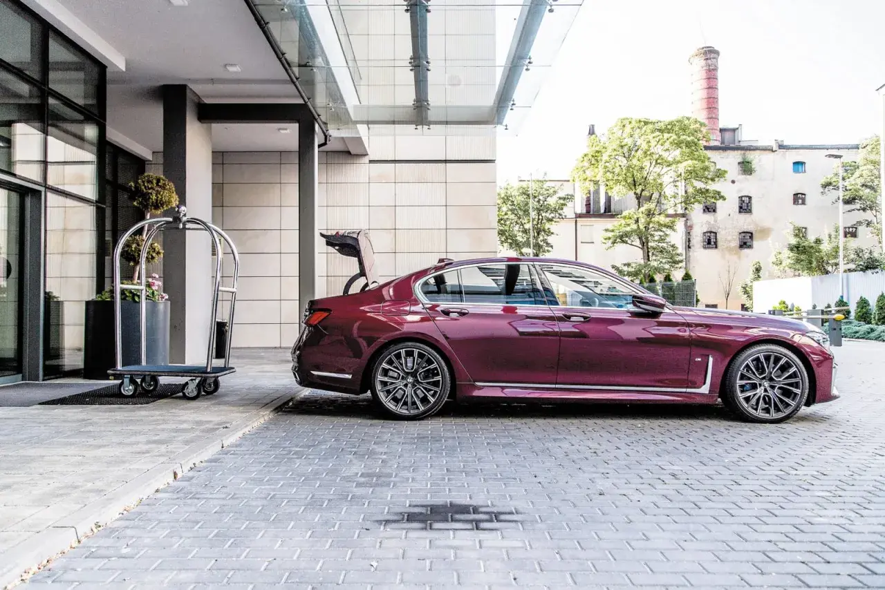 Eleganckie bmw g11 w kolorze śliwkowym z otwartym bagażnikiem stoi przed hotelem obok wózka na bagaż.