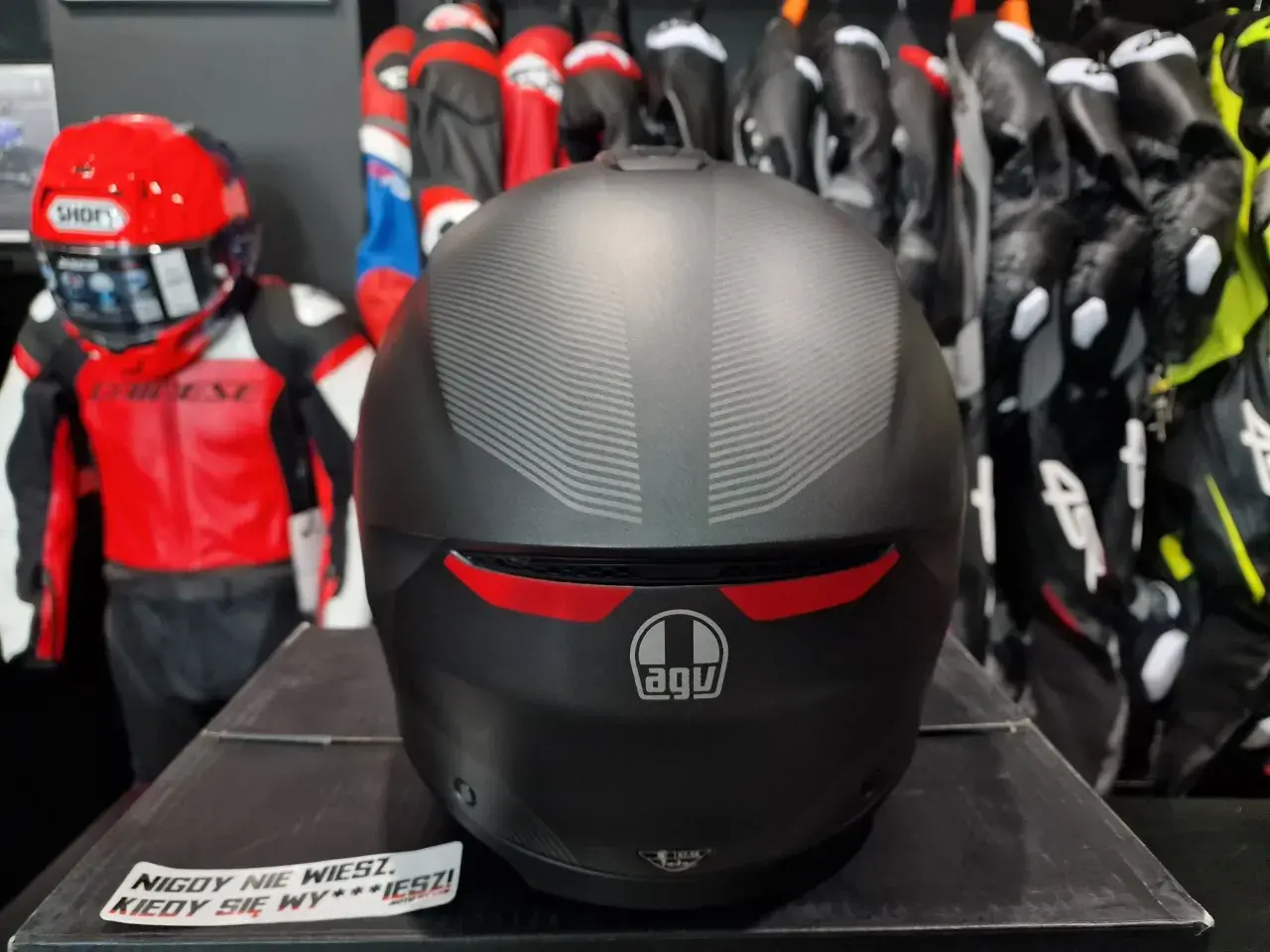 Matowy kask AGV z subtelnym wzorem, gotowy na przygodę. W tle czerwono-czarny kombinezon motocyklowy.