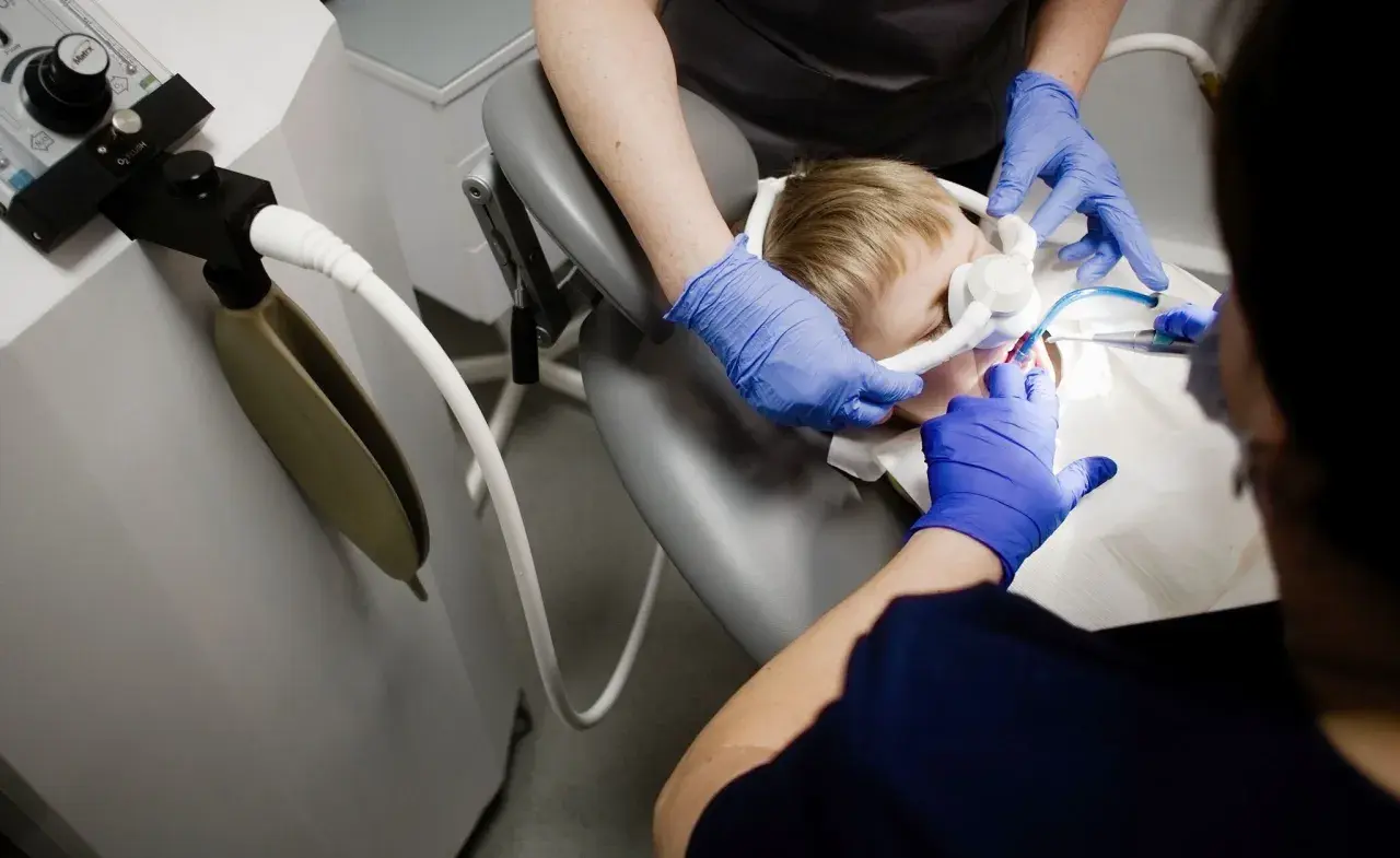 Chłopiec w masce do znieczulenia ogólnego u dentysty. Dwie osoby w niebieskich rękawiczkach wykonują zabieg.
