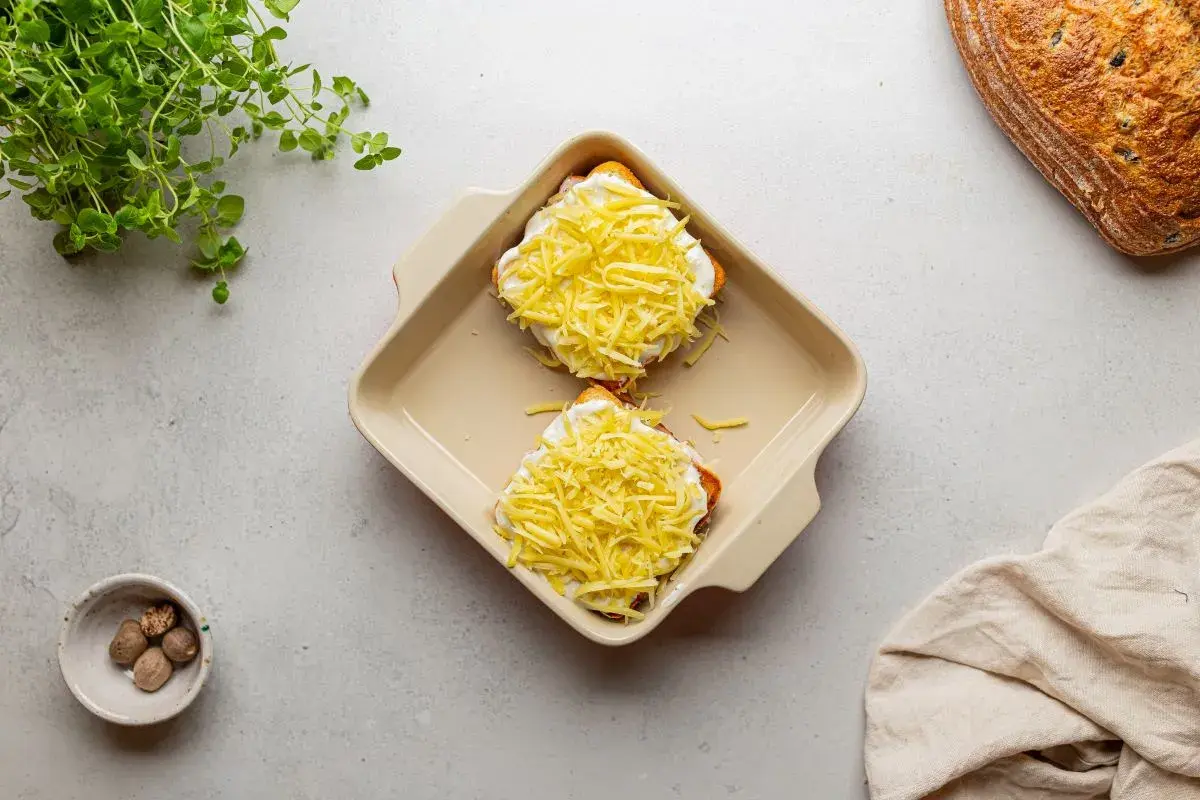 Dwa tosty z szynką i serem, przygotowane do zapieczenia, jak w klasycznym croque madame. Obok gałka muszkatołowa i świeża zielenina.