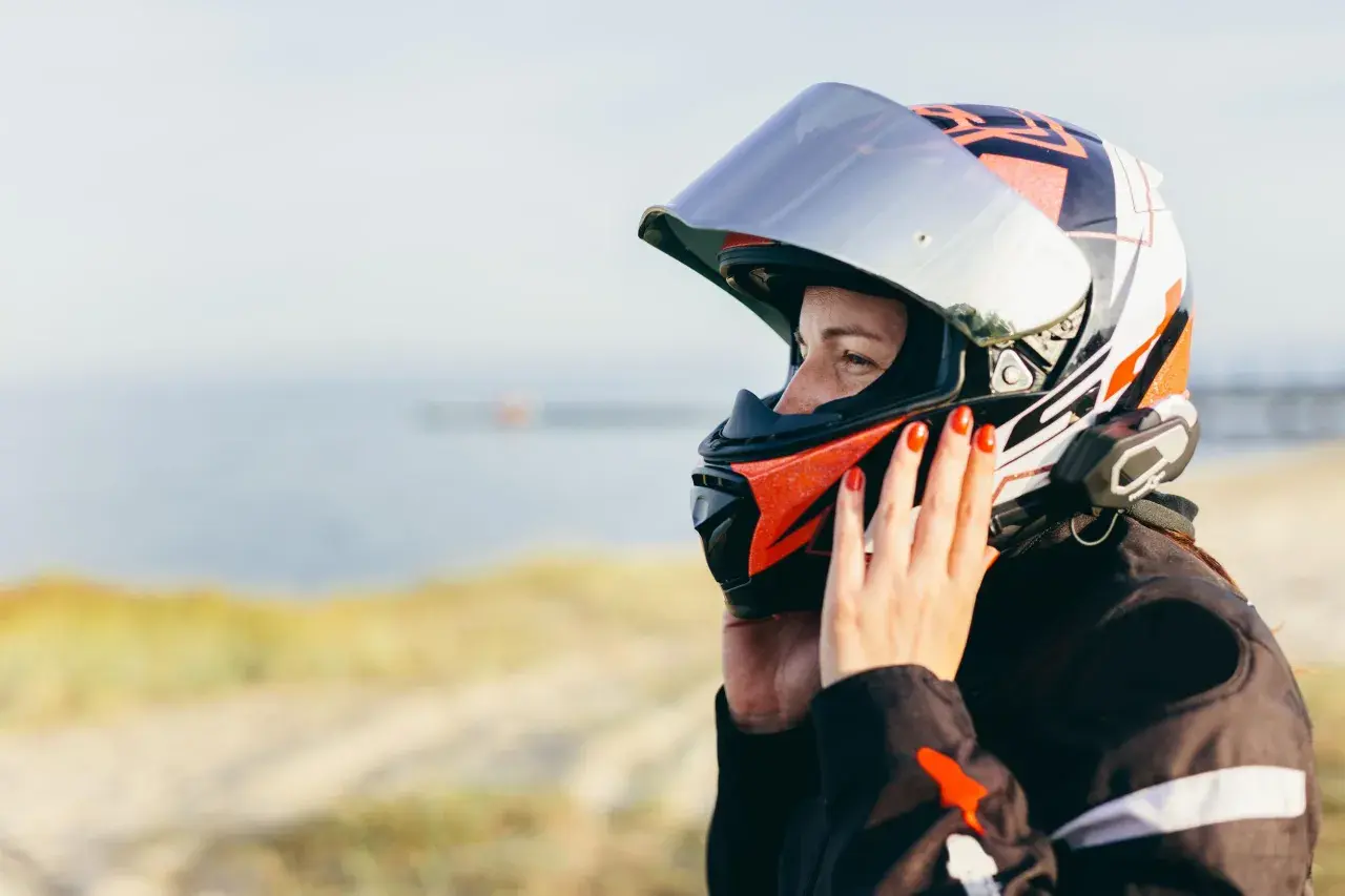 Donna con casco da moto colorato, con mani che aggiustano la visiera per evitare che si appanni. Sfondo sfocato di mare e sabbia.