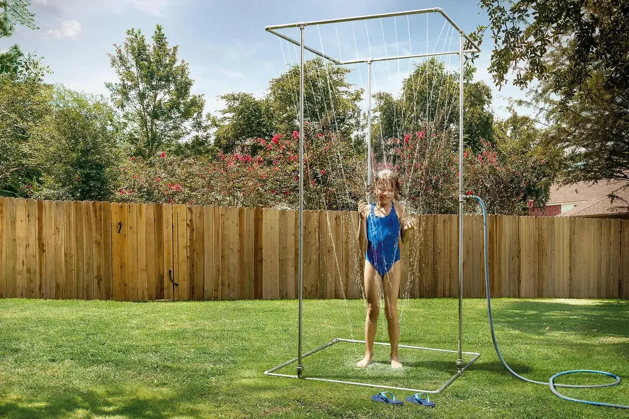 Ein Mädchen im blauen Badeanzug genießt eine erfrischende Dusche unter einer selbstgebauten Gartendusche im sonnigen Garten.