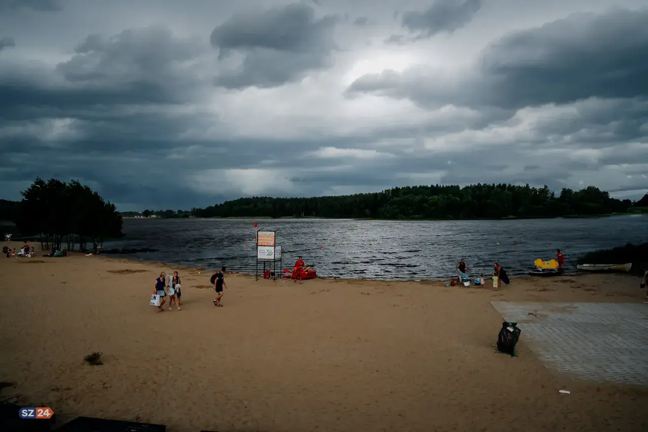 Plaża nad jeziorem Wysoka, z ciemnymi chmurami na niebie. Ludzie spacerują po piasku, niektórzy odpoczywają. W tle las i spokojna tafla wody.