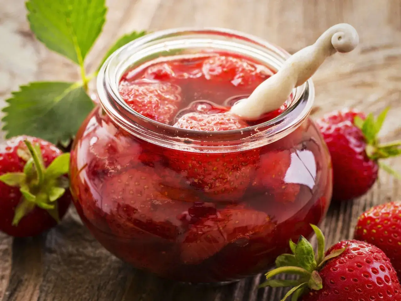 Ein Glas mit ganzen Erdbeeren und Sirup, bereit zum Erdbeeren einwecken. Frische Erdbeeren und Blätter liegen daneben.