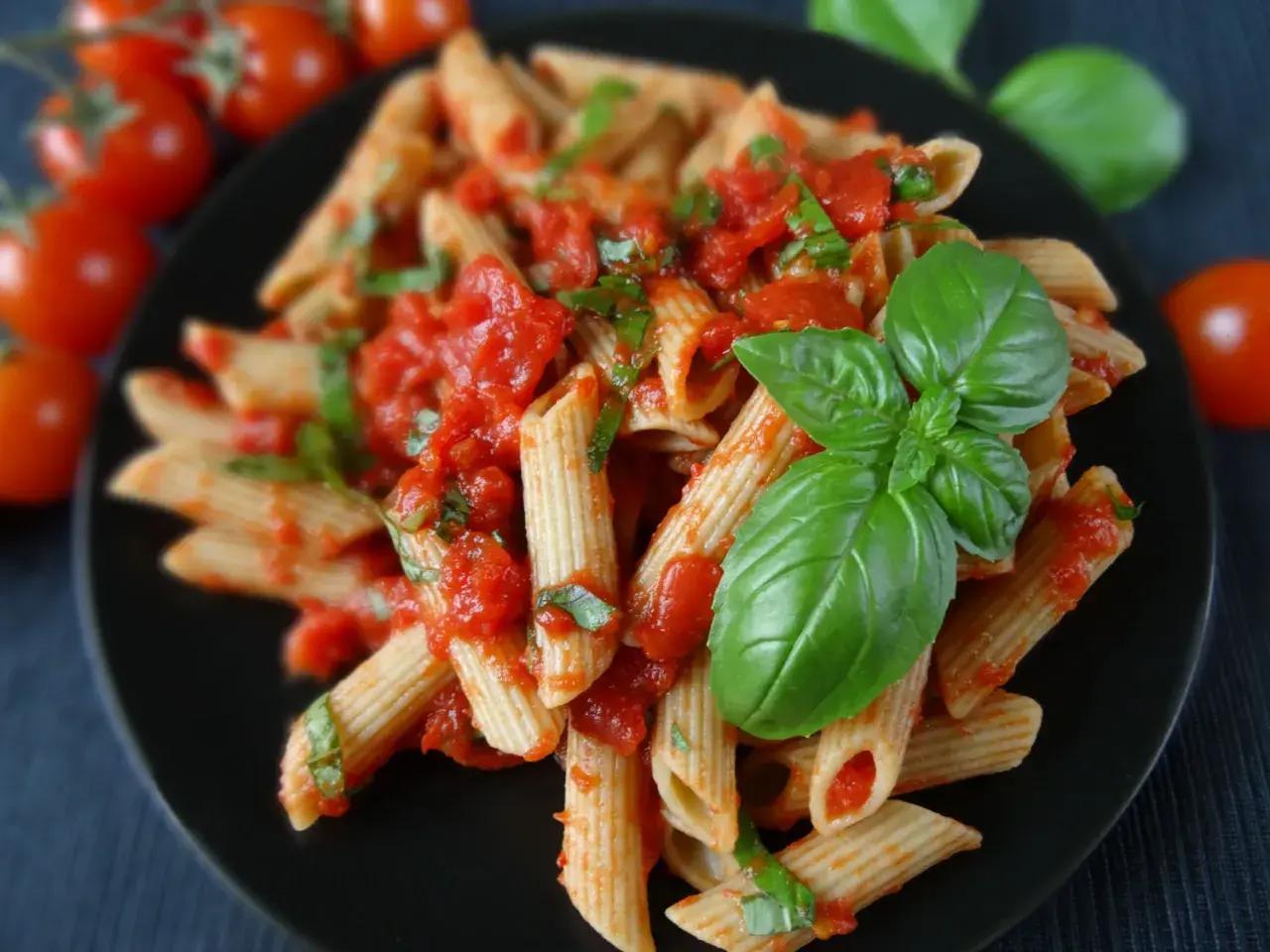Penne z pomidorami i świeżą bazylią, polane aromatycznym **sosem bazyliowym do makaronu**. Obok gałązka pomidorków koktajlowych.