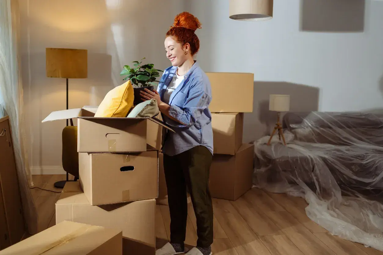 Donna con pianta in mano, circondata da scatoloni, in una casa in fase di trasloco. Potrebbe essere interessata alle agevolazioni prima casa senza residenza.