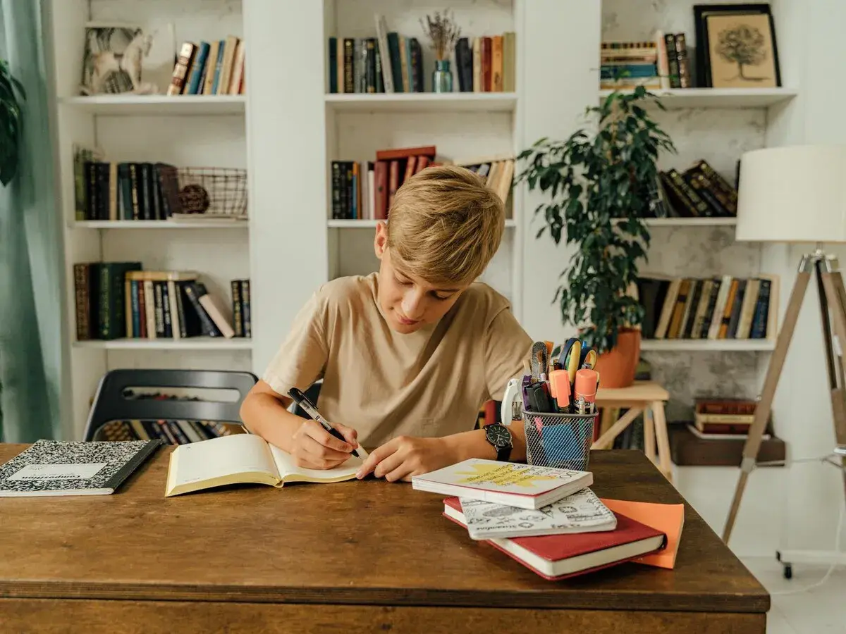 Un niño concentrado escribe en su cuaderno, rodeado de libros y útiles escolares, practicando dictados cortos para mejorar su ortografía.