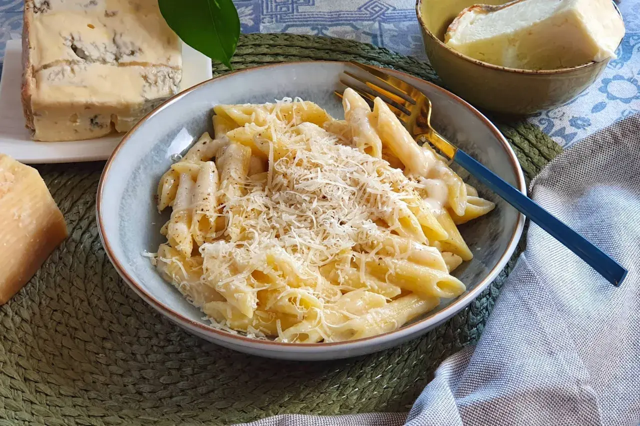 Piatto di penne cremose, condite con una ricca salsa di spaghetti ai 4 formaggi, guarnite con parmigiano grattugiato. Accanto, pezzi di formaggio fresco e stagionato.