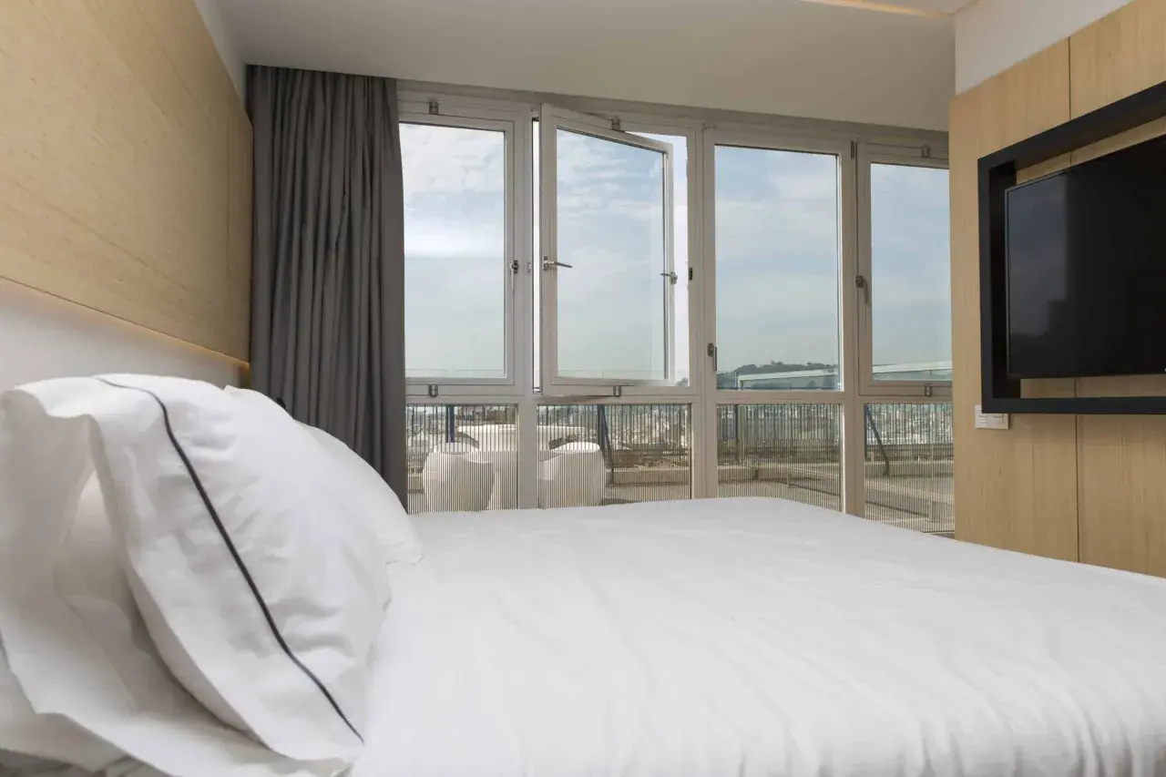 Cama blanca con almohadas, cortinas grises y ventanas de triple cristal que dejan ver el cielo y la ciudad.