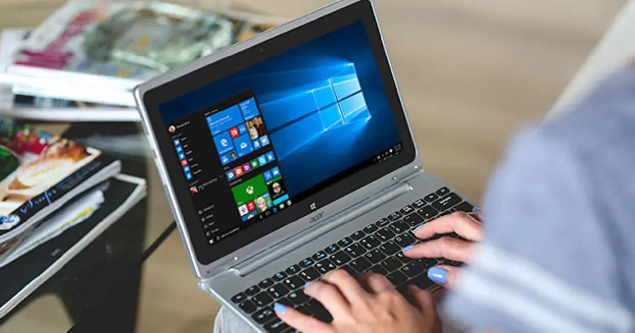 Kobieta pisze na laptopie z systemem Windows 10. W tle widać stosy czasopism, co sugeruje, że zadania w tle ochrony systemu są wykonywane podczas relaksu.