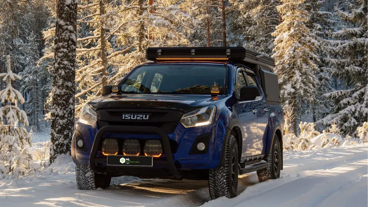 Niebieski Isuzu w zimowym lesie, z dodatkowymi światłami LED na dachu i zderzaku. Dowiedz się, jak ustawić żarówki LED w samochodzie, by podróżować bezpiecznie.