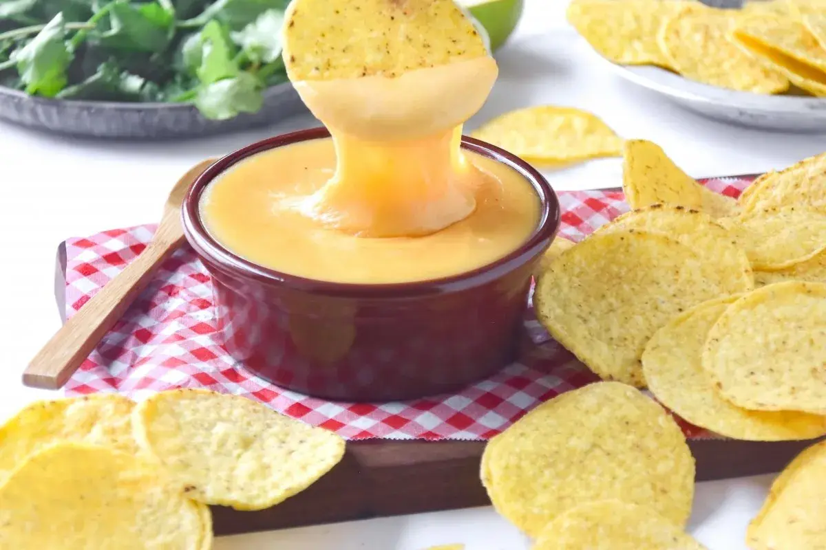 Un totopo se sumerge en una cremosa salsa cheddar, rodeado de más totopos y cilantro fresco.