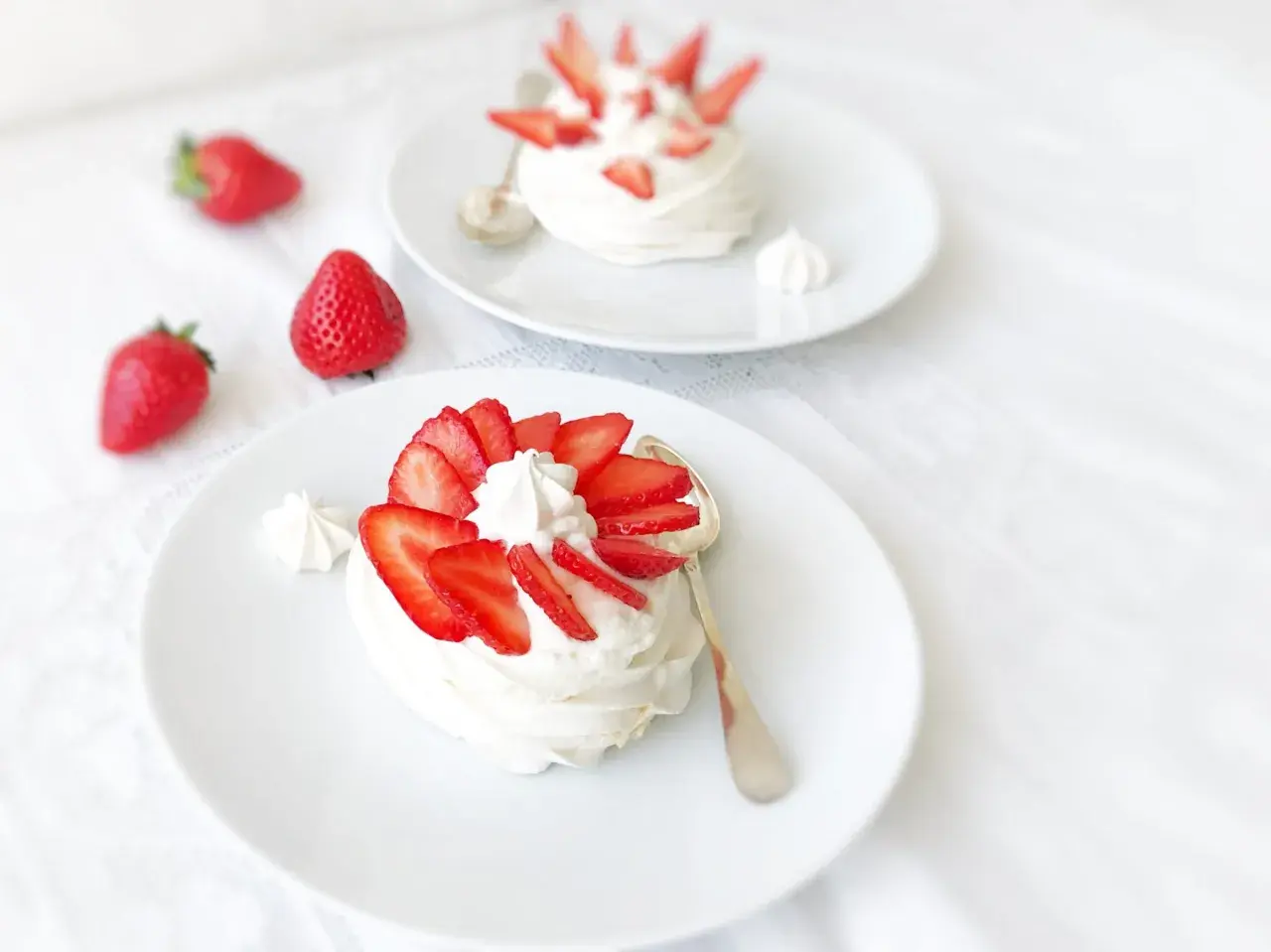 Deux pavlovas à la fraise, légers et aériens, ornés de fruits rouges frais et de crème fouettée, sur des assiettes blanches.