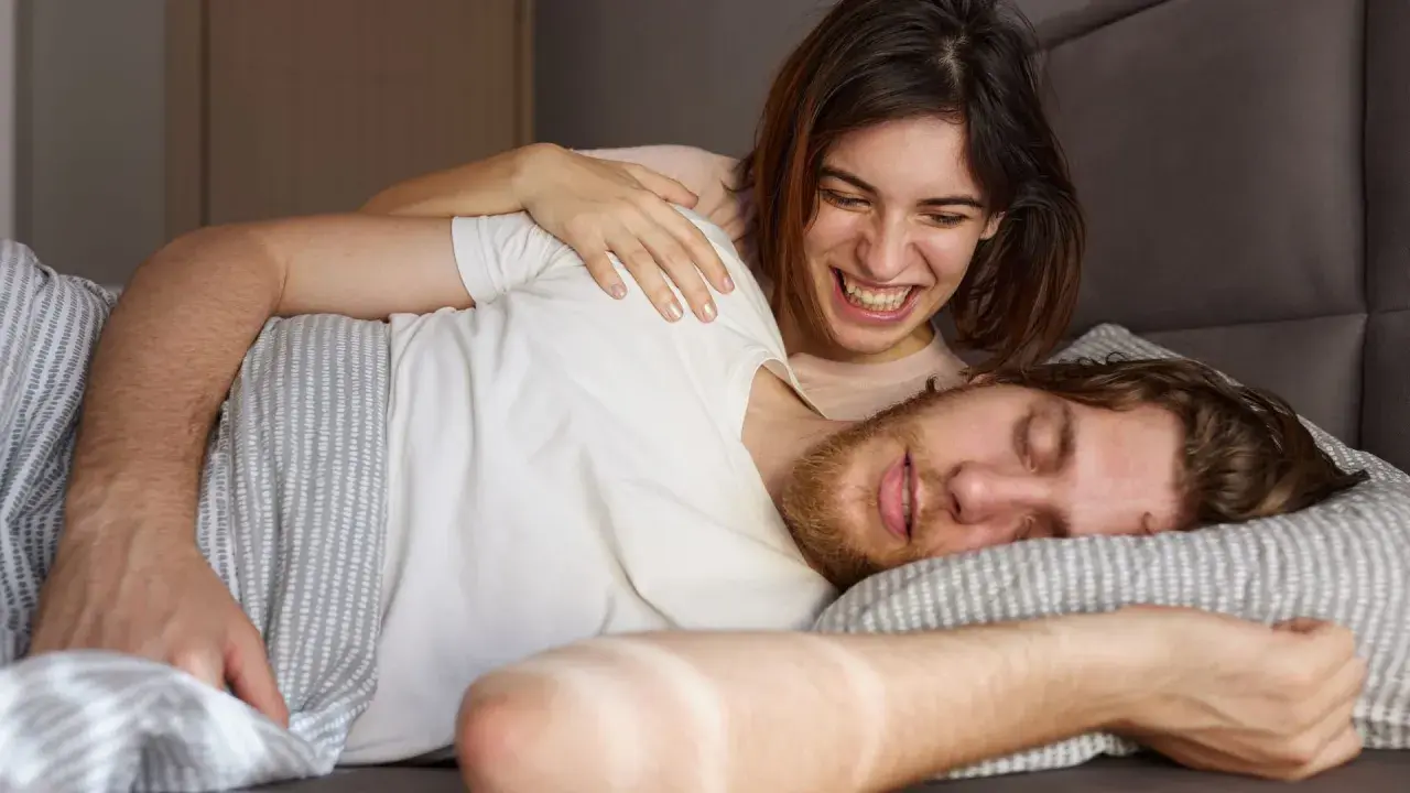 Ein Mann schläft, während eine Frau ihn lachend kitzelt. Vielleicht redet er im Schlaf und sie amüsiert sich.