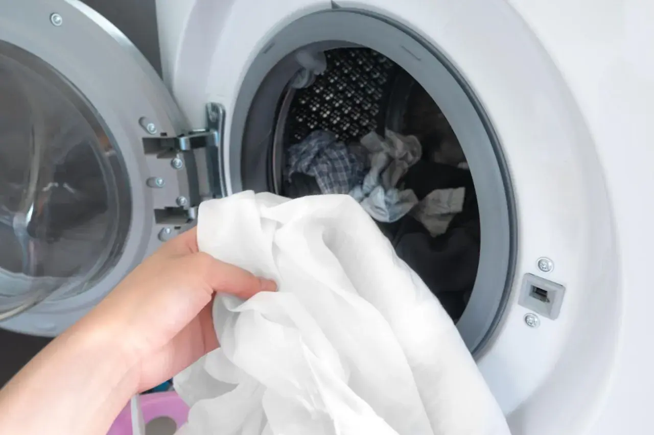 Eine Hand hält ein weißes Hemd, bereit, es in die Waschmaschine zu legen. Die Tür der Waschmaschine ist geöffnet und gibt den Blick auf weitere Wäsche frei.